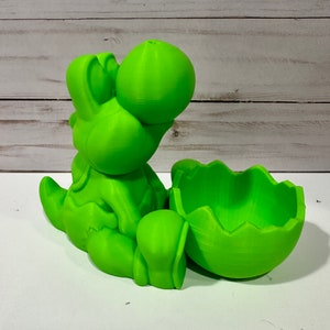 May include: A green 3D printed Yoshi figurine with an open egg shell. The Yoshi is sitting with its legs crossed and its head tilted back. The egg shell is open and has a rough texture.