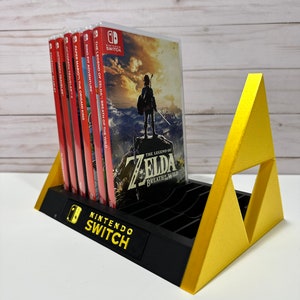 Może przedstawiać: Czarno-złoty stojak na gry Nintendo Switch o trójkątnym kształcie. Stojak mieści wiele kartridży z grami i zawiera logo Nintendo Switch. Stojak prezentuje kartridż z grą The Legend of Zelda: Breath of the Wild.