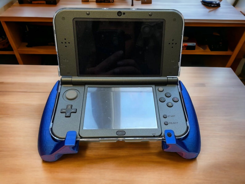 K&ouml;nnte beinhalten: Eine schwarze Nintendo 3DS Handheld-Spielekonsole mit blauen Griffaufs&auml;tzen. Die Konsole ist ge&ouml;ffnet und der obere Bildschirm zeigt einen schwarzen Bildschirm an. Der untere Bildschirm zeigt einen grauen Bildschirm an. Die Konsole verf&uuml;gt &uuml;ber verschiedene Tasten, darunter eine Starttaste, eine Select-Taste und eine Power-Taste.