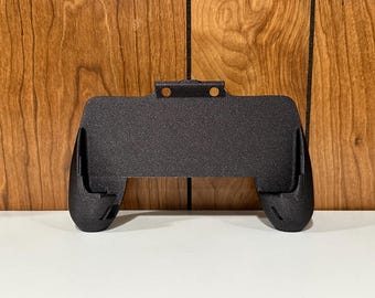 Peut inclure: Support de manette de jeu noir et texturé, conçu pour maintenir un appareil de jeu. Le support a un centre rectangulaire avec deux poignées incurvées de chaque côté. Il comporte un support de montage avec deux trous en haut. Le fond est un mur en bois.