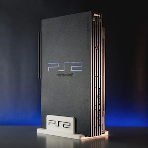 Peut inclure: Une console Sony PlayStation 2 noire avec un support blanc. La console a une façade argentée avec le logo PlayStation 2 et le texte "PlayStation 2".