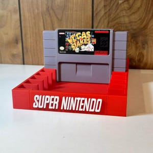 May include: A red plastic Super Nintendo game cartridge holder with a grey Super Nintendo game cartridge inside. The game cartridge is for the game "Vegas Stakes".