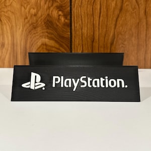 Peut inclure: Support noir avec le logo PlayStation et lettrage blanc. Le support présente le logo PlayStation et le mot "PlayStation" dans une police de caractères nette et audacieuse. Conçu pour une console de jeu.