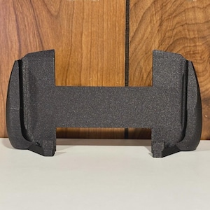 May include: A black, textured, 3D-printed object with a unique shape. It has a rectangular center section with curved, rounded ends. The object is designed to hold something, with a slot in the center. The background is a wooden surface.