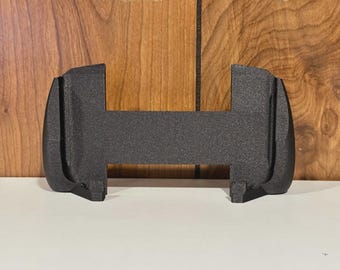 Peut inclure: Un objet noir texturé, imprimé en 3D, de forme unique. Il possède une section centrale rectangulaire avec des extrémités incurvées et arrondies. L'objet est conçu pour contenir quelque chose, avec une fente au centre. Le fond est en bois.