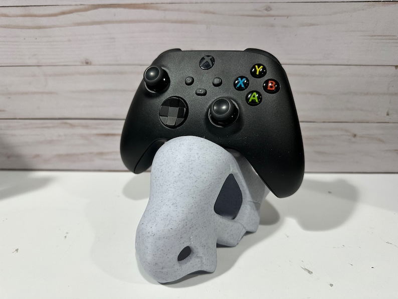May include: A black Xbox controller with colored buttons sits on a white, speckled, 3D printed stand.