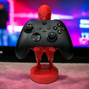 May include: A red and black video game controller stand shaped like a superhero. The controller stand is holding a black Xbox controller.