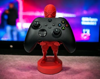 May include: A red and black video game controller stand shaped like a superhero. The controller stand is holding a black Xbox controller.