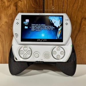 Op de afbeelding: Witte Sony PSP handheld gameconsole met een zwarte grip bevestigd. Het scherm toont een videomenu met tekst. De console heeft een D-pad, actieknoppen en een startknop. De PSP rust op een houten oppervlak.