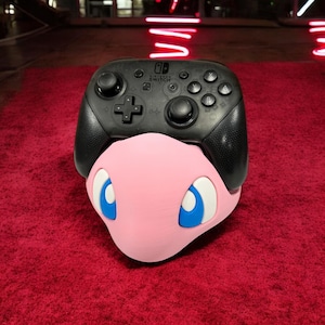 May include: A black Nintendo Switch controller with a pink and blue Jigglypuff character stand. The controller has a black and white Nintendo Switch logo on the front.
