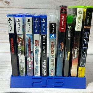 May include: A blue plastic video game case holder with a PS5 logo on the front. The holder is filled with video game cases for various consoles, including PS5, PS4, Xbox 360, Xbox One, and PlayStation 2.