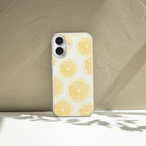 May include: A white phone case featuring a pattern of yellow lemon slices. The case has a clear border and a camera cutout. The background is a textured beige wall and a white surface with shadows.