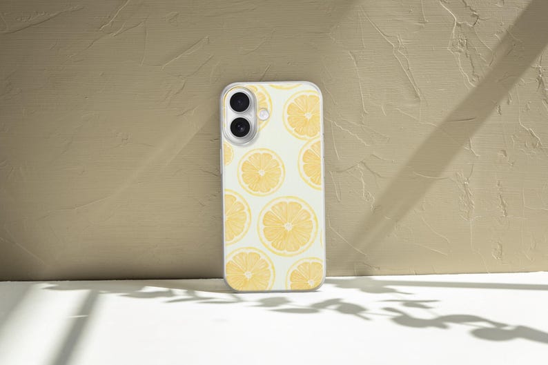 May include: A white phone case featuring a pattern of yellow lemon slices. The case has a clear border and a camera cutout. The background is a textured beige wall and a white surface with shadows.