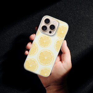 May include: A phone case featuring a repeating pattern of yellow lemon slices on a light background. The case is held in a hand, showcasing the design and the phone's camera array.