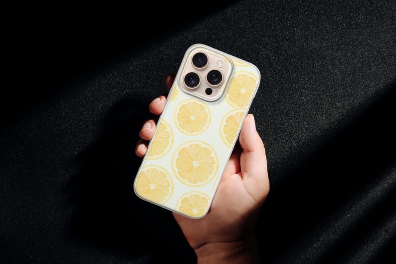 May include: A phone case featuring a repeating pattern of yellow lemon slices on a light background. The case is held in a hand, showcasing the design and the phone's camera array.