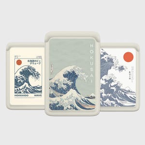Puede incluir: Tres fundas blancas para teléfono con arte de olas japonesas. La funda central presenta una gran ola con la palabra "HOKUSAI" verticalmente. Las otras fundas tienen diseños de olas y sol con texto japonés. Las fundas están en un soporte gris claro.