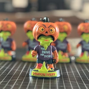 May include: A collection of Halloween-themed bobblehead figurines. Each figurine features a pumpkin head with a green, vomit-like substance, wearing a purple shirt with the text "Too Ghoul for Skool". The base reads "Pumpkin Pukey".