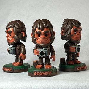 May include: Three brown Bigfoot figurines holding cameras. Each figure has a brown face, hair, and body, with a gray camera. The figurines stand on a green base with the word "STOMPZ" in brown. The Bigfoot is smiling.