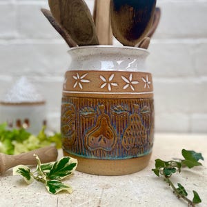 May include: A ceramic utensil holder with a white top and a brown band featuring white flower cutouts. The lower portion has embossed fruit designs. Wooden spoons and spatulas are inside the holder.