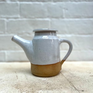 May include: A handcrafted ceramic teapot, featuring a light grey upper section and a tan base. It has a curved spout, a handle, and a lid. The background is a white brick wall.