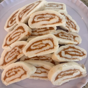 May include: A pile of sliced, spiral-shaped pastries with a white exterior and a tan, swirled filling. The pastries are arranged on a white plate with a scalloped edge, creating a visually appealing presentation of the baked goods.