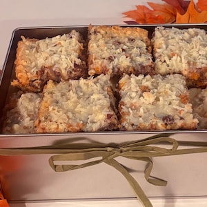 May include: A rectangular silver metal tin filled with square baked bars topped with shredded coconut. The tin is tied with a thin green ribbon and surrounded by decorative orange, white, and yellow pumpkins and autumn-colored leaves.