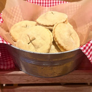 May include: A round metal basin filled with several small, golden-brown, circular hand pies. The pies feature a crimped edge and a small cross-shaped vent in the center. The basin is lined with light brown parchment paper and a red and white gingham cloth.