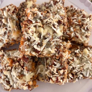 May include: A plate of homemade coconut pecan bars. The bars are square-shaped, with a base of pecans and a white, creamy layer topped with shredded coconut. The bars are arranged on a white plate.