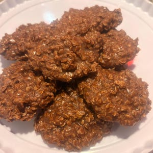 May include: A close-up shot of a white plate holding several chocolate no-bake cookies. The cookies are a rich brown color, with a textured surface from the oats. The cookies are arranged in a slightly overlapping pattern.