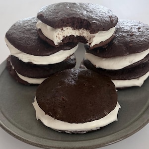 May include: A stack of round chocolate sandwich cookies with white cream filling on a gray plate. The top cookie has a visible bite mark, revealing the dark brown cake-like texture and the thick layer of white frosting between the two halves.
