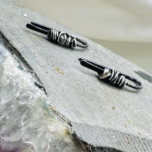 May include: Two silver-toned barrette hair clips with a dark, possibly black, central element. The clips are wrapped with silver wire and rest on a gray, textured surface. The background is a light, neutral color.
