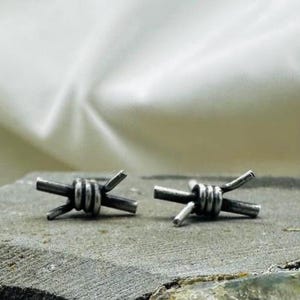 Puede incluir: Un par de pendientes de botón plateados con forma de alambre de púas en miniatura. Los pendientes están detallados con alambre enrollado y púas afiladas y puntiagudas. Se exhiben sobre una superficie gris texturizada.