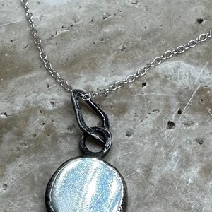 May include: A silver necklace with a round pendant. The pendant is made of metal and has a textured surface. The chain is silver and has a delicate design.