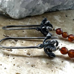 May include: A pair of silver-toned earrings. Each earring features a floral design and a series of faceted, reddish-brown beads. The earrings are displayed on a textured, light-colored surface with a gray stone in the background.