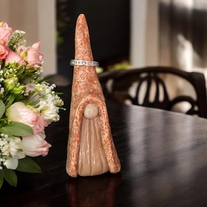 May include: A ceramic gnome ring holder with a textured, reddish-brown finish. A silver ring with small, clear stones is placed on the gnome's pointed hat. The gnome has a beige face and body. A bouquet of pink roses and white flowers is in the background.