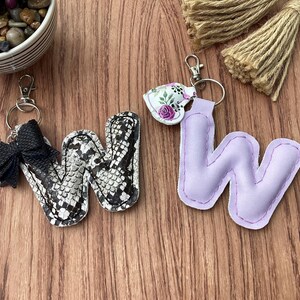 May include: Two keychains shaped like the letter "W". One is black and white snakeskin print, the other is lavender with a floral accent. Both have silver-tone hardware and key rings. The background is a wooden surface.