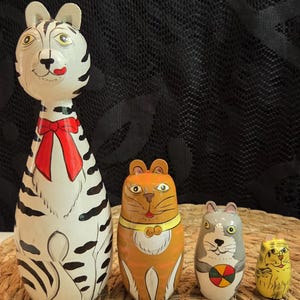 Könnte beinhalten: Ein Set aus vier handbemalten hölzernen Matroschka-Puppen in Katzenform. Die größte ist weiß mit schwarzen Streifen und einer roten Schleife. Die zweite ist orange mit einem gelben Halsband. Die dritte ist grau und hält einen bunten Ball.