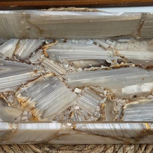 Peut inclure: Un plateau rectangulaire composé de morceaux polis de pierre d'agate translucide grise et blanche. Le plateau présente des parois hautes et une base plate, avec des bandes naturelles visibles et des inclusions minérales brun doré.