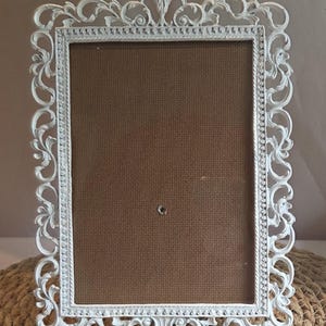 May include: A rectangular picture frame featuring an ornate white border with intricate scrollwork and floral detailing. The frame surrounds a solid brown, textured backing material that includes a single small circular hole in the center.