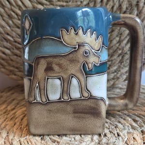 May include: A square-shaped ceramic mug featuring a relief design of a brown moose standing on a tan ground against a blue sky background. The mug has a thick, rectangular handle and a smooth, glazed finish with visible color transitions.