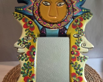 May include: A rectangular wall mirror featuring a hand-painted frame with a sun face at the top. The frame is decorated with yellow, red, and green floral patterns and two side faces. The mirror surface has a textured, etched leaf-like design.