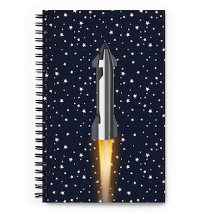 May include: Spiral-bound notebook featuring a rocket ship blasting off against a dark blue starry sky background. The rocket is silver and white with orange flames. The notebook has a black spiral binding.