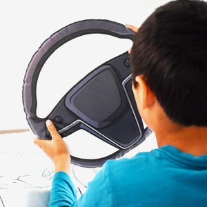 May include: A gray and black steering wheel-shaped pillow. The pillow has a cartoon-like design with a dark gray rim and a black center. The pillow is being held up by a person wearing a blue shirt.