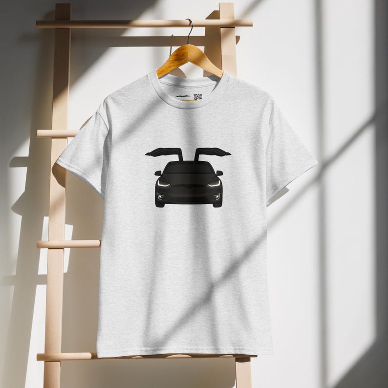 May include: A light gray t-shirt featuring a black illustration of a car with its doors open. The car's headlights and front are visible. The shirt is hanging on a wooden hanger.