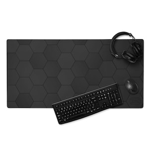 Puede incluir: Alfombrilla de escritorio negra con un patrón hexagonal, un teclado, un ratón y unos auriculares negros. La alfombrilla proporciona una gran superficie para una configuración informática. El teclado está inclinado hacia la parte inferior de la imagen.