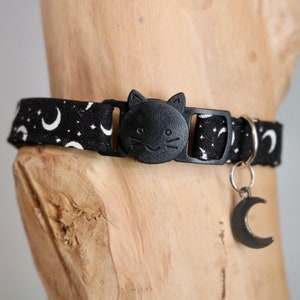 May include: Black cat collar with a white crescent moon and star pattern. The collar has a black cat face buckle and a silver crescent moon charm.