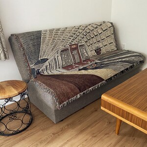 K&ouml;nnte beinhalten: Ein graues Sofa mit einer gewebten Wanddecke, die eine U-Bahn-Szene zeigt. Die Decke hat eine braune, graue und wei&szlig;e Farbgebung. Das Sofa steht in einem Raum mit Holzboden und einem Holz-Couchtisch.