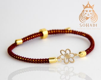 May include: A red string bracelet with gold accents and a gold flower charm. The bracelet is made of a braided cord and has a small gold bead at the end. The flower charm is delicate and intricate.