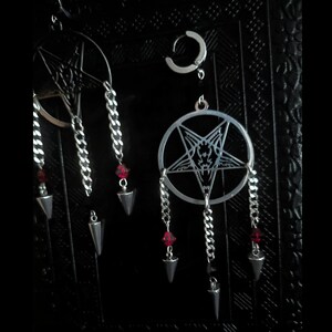 May include: Silver-toned earrings with a gothic theme. The earrings feature a pentagram design, chains, red beads, and silver-colored arrow-shaped pendants. A small hoop earring is also visible. The background is dark.