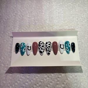 May include: A collection of ten press-on nails with diverse designs. The nails showcase patterns like cow print, paisley, and a horseshoe. Colours include black, white, teal, and mauve. The nails are presented on a white card.